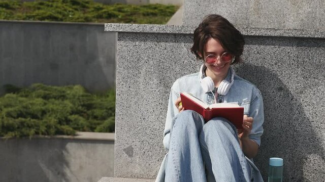 Young calm happy woman 20s wear denim shirt pink t-shirt headphones on neck read book rest relax in city sit on concrete stairs sunshine day outdoor nature. Urban lifestyle leisure sun light concept