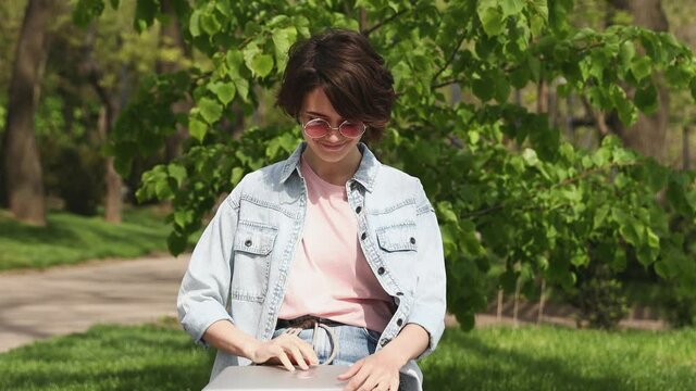 Young woman 20s wear denim shirt pink t-shirt glasses user work on laptop pc computer sit on bench rest in spring green city park sunshine day outdoors nature. Mobile office freelance business concept