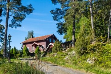 Fototapeta na wymiar Traditional Swedish wooden house near forest. Countryside village in Scandinavia. Red typical cottage rural house with porch. Carport. In courtyard Landscaping of garden with ornamental thuja plants.