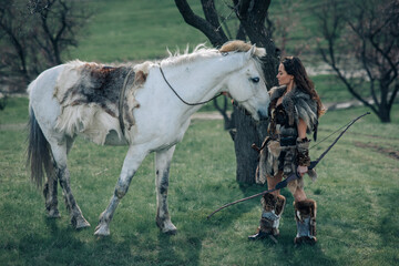 Woman stands in image of warrior amazon with bow in her hands near her horse.