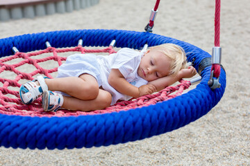 Little toddler boy, sleeping in a round swing on a playground, tired after full day of games