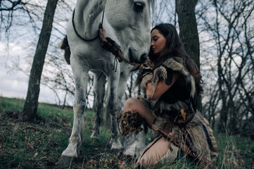 Woman sits and embraces her horse in forest in image of warrior amazon.