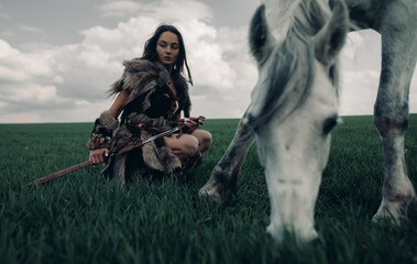 Woman sits on meadow in image of warrior amazon near grazing horse.