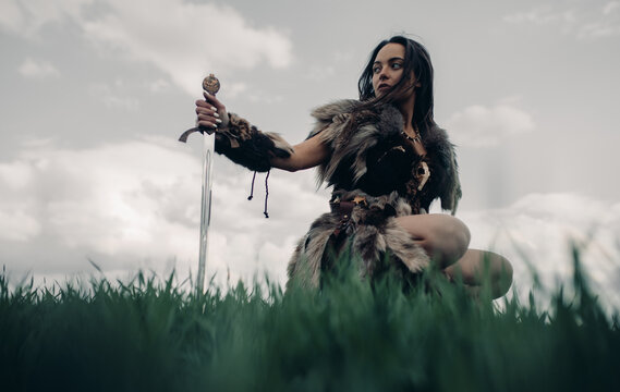 Woman sits on meadow in image of warrior amazon with sword in her hand.
