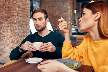 woman having dinner at a table in a cafe and a man with a cup of coffee in the background