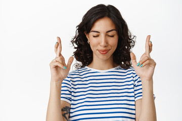 Image of happy optimistic girl making wish, close eyes and cross fingers while anticipating, praying for good results, standing hopeful against white background