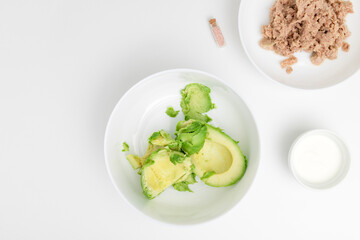 Peeled avocado on the white plate. Yogurt and tuna are placed near. Flat lay. Step-by-step recipe.