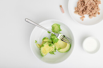 Peeled avocado on the white plate with fork. Yogurt and tuna are placed near. Flat lay. Step-by-step recipe.