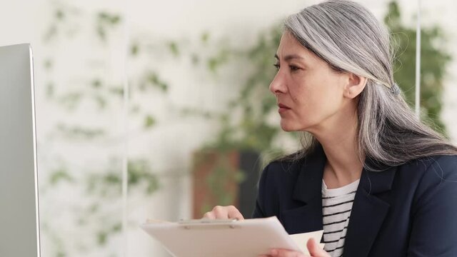 Side view of an Asian confident woman with gray hair checking papers and looking at the monitor while sitting at a table in the office