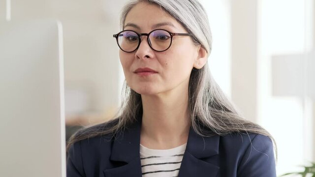A confident asian woman with gray hair and eyeglasses looking at the monitor and then typing on the keyboard while sitting at a table in the office
