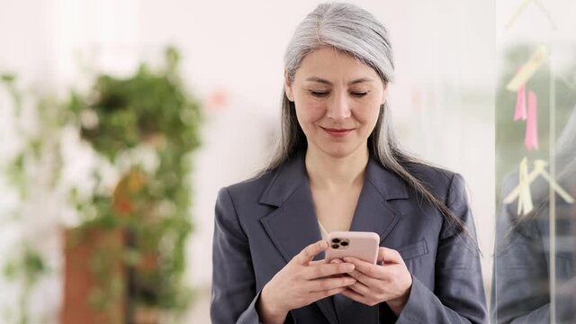 A positive asian woman in a suit with gray hair leafing something on the phone and looking to the side in the office