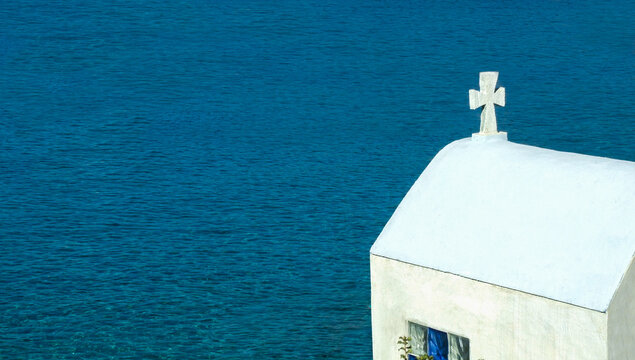 Little white church, traditional Greek chapel against the turquoise sea and blue cloudy sky.