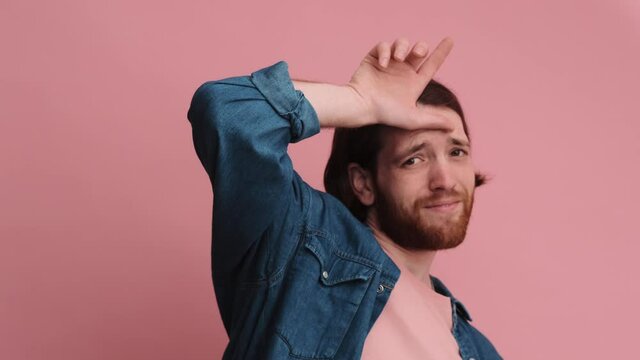 A ridiculous man is showing looser sign abusing fooling grimacing isolated on a pink color background