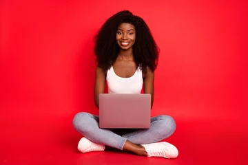 Poster Im Rahmen Portrait of beautiful trendy cheerful girl sitting lotus pose using laptop study learn isolated over bright red color background © deagreez