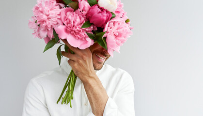 Closeup of handsome man covers his eyes with the bouquet of pink peonies as a gift for Valentine's day or wedding day. Smiling male carrying flowers in the hands, isolated on the grey background.