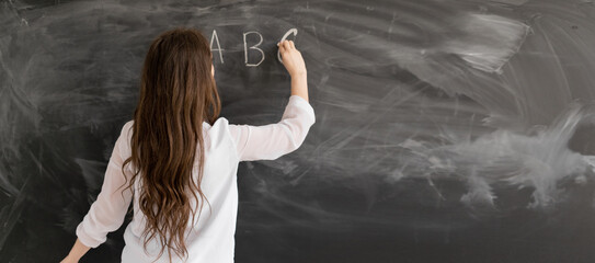 Rear view of a student or teacher with long brunette hair writing on a blank chalk board or...