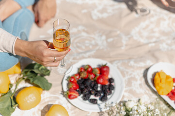 Close-up woman hand holding a glass of white wine sitting on a blanket picnic with fruits.
