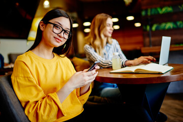 Asian woman browsing smartphone while female friend working on laptop in cafeteria