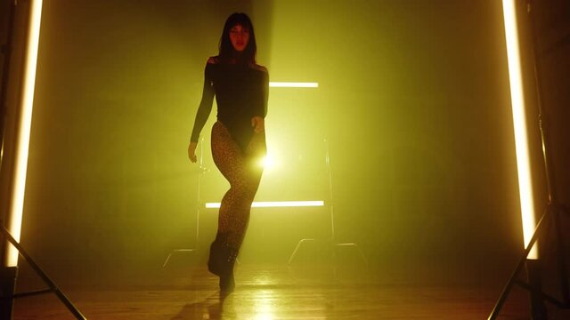 Amazing woman walking between long lamps and looks seductively into camera. Sexy outfit - heels, bodysuit and leopard pantyhose. Model struts down runway .