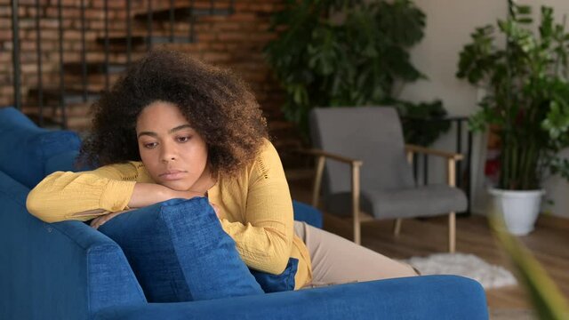 Depression concept. Upset African-American woman feels bored sitting on the couch at home alone, desperate black lady feels lonely, missing boyfriend, multiracial female worried about future