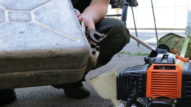 the lawnmower pours gasoline from a metal can into the mower container through a plastic funnel, refueling the lawnmower with fuel, pouring gasoline into the mower by a person