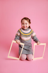 Portrait of a little smiling girl sitting and holding two blank frames for mock-up on a pink...