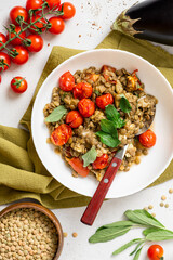 Stewed green lentils with tomatoes, eggplants, spices, and herbs in a white plate on a gray concrete background top view. Vegan food. Lentils with vegetables.