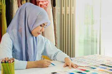 Portrait of beautiful asian muslim woman designer with hijab drawing and design dress in her home working studio room with sample color cloth, dress.