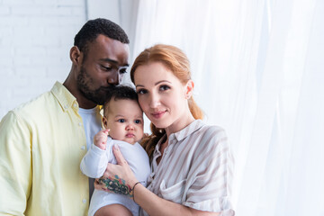 happy woman looking at camera near smiling husband kissing head of infant daughter