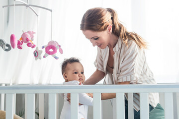happy woman holding african american kid near crib and baby mobile with toys
