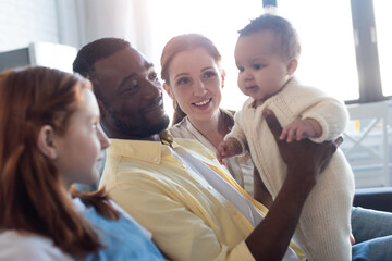smiling african american father holding infant child near blurred family