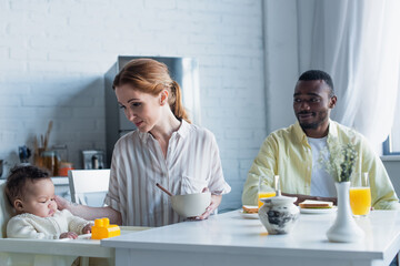 woman holding bowl near baby girl and smiling african american husband in kitchen