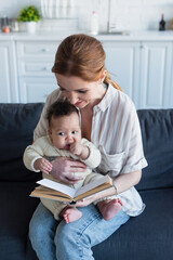 Fototapeta premium woman reading book while sitting on sofa with infant african american kid