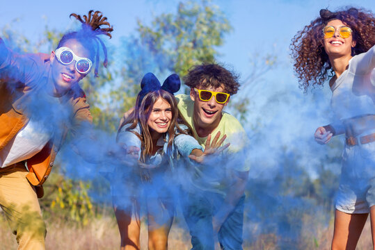 Beautiful young man and woman hold light up colored smoke bombs - Happy friends having fun in the park with multicolored smoke bombs - Young students celebrating spring break together. Holi festival.