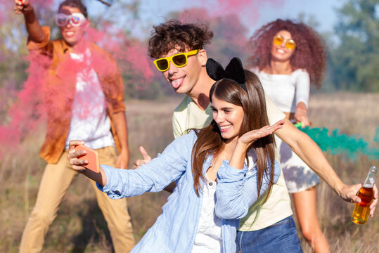 Beautiful young woman hold smartphone, doing selfie with happy friends, having fun in the park with multicolored smoke bombs smoke fog. Young students celebrating spring break together. Holi festival.