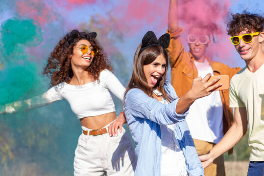 Beautiful young woman hold smartphone, doing selfie with happy friends, having fun in the park with multicolored smoke bombs smoke fog. Young students celebrating spring break together. Holi festival.
