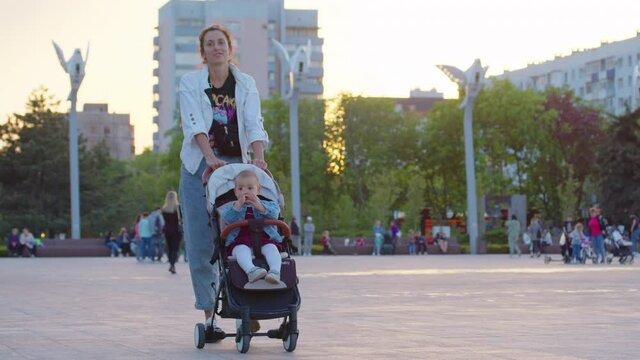 Caucasian woman walking along the alley in summer park with baby stroller. Portrait of confident young mother strolling with infant in baby carriage outdoors.