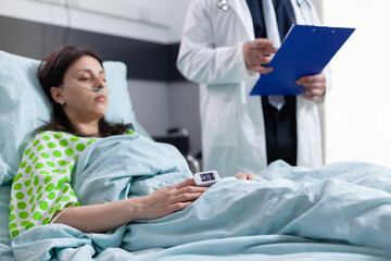 Close up hospitalized woman with finger heart rate monitor showing pulse while resting in bed during cardiology consultation in hospital ward. Patient with oximeter waiting for sickness treatment
