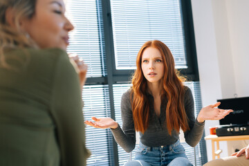 Portrait of dumbfounded young redhead woman looks away with hands spread in incomprehension during brainstorming of startup projects in modern office room near window.