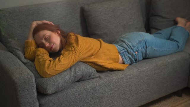 Peaceful smiling redhead female woman with serene face relax at home lying down in comfortable sofa couch enjoy welfare peace of mind. Happy pretty young woman sleeping on stomach in cozy living room.