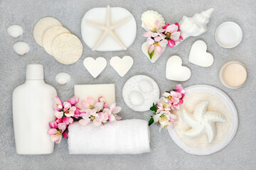 Natural skincare cleansing ingredients & products with soaps, scrubs, sponges, moisturising cream & apple blossom flowers with decorative seashells. Flat lay on mottled grey background.