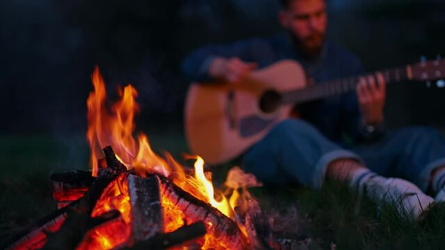 Man sitting by bonfire with guitar. Lonely man sitting by bonfire on the beach near forest playing acoustic guitar