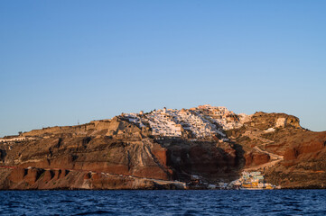 Fototapeta na wymiar View from sea of Oia up the hill and sea port. Santorini, Greece. White city architecture. Luxury tourism. Sunset light.