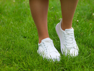 Legs of a girl in white sneakers on green grass (lawn). Close up of a pair of white shoes on the legs of a girl against a background of green grass.