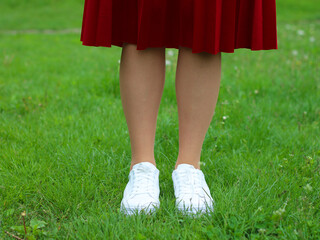 Legs of a girl in white sneakers on green grass (lawn). Close up of a pair of white shoes on the legs of a girl against a background of green grass.