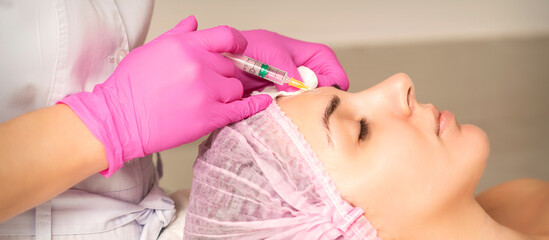 Obraz na płótnie Canvas Young woman receiving an injection of anti-aging botox filler to the forehead from a cosmetologist in a beauty salon. Facial treatment injection