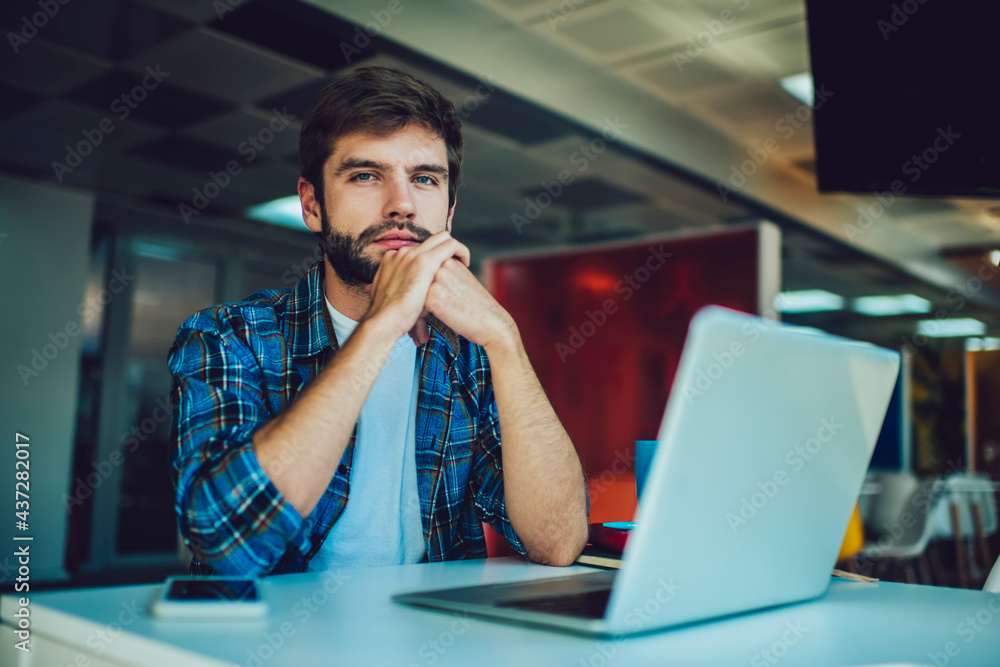 Poster portrait of caucasian hipster guy with netbook looking at camera during distance job in coworking sp
