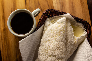 Duas tapiocas dentro de uma pequena cesta de vime e uma xícara de café sobre mesa de madeira.