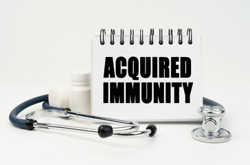 On a white table lies a stethoscope, pills and a notebook with the inscription - ACQUIRED IMMUNITY