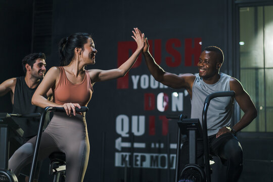 Asian female and african male sportsperson giving high five together after training on stationary bicycle. Health care and fitness in gym concept